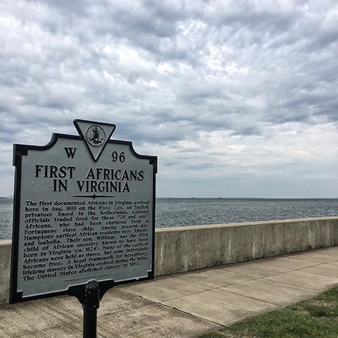 A sign explains how the first Africans in arrived Virginia. It stands along a sidewalk next to the water.