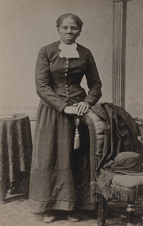 Harriet Tubman stands in a black dress next to a chair.
