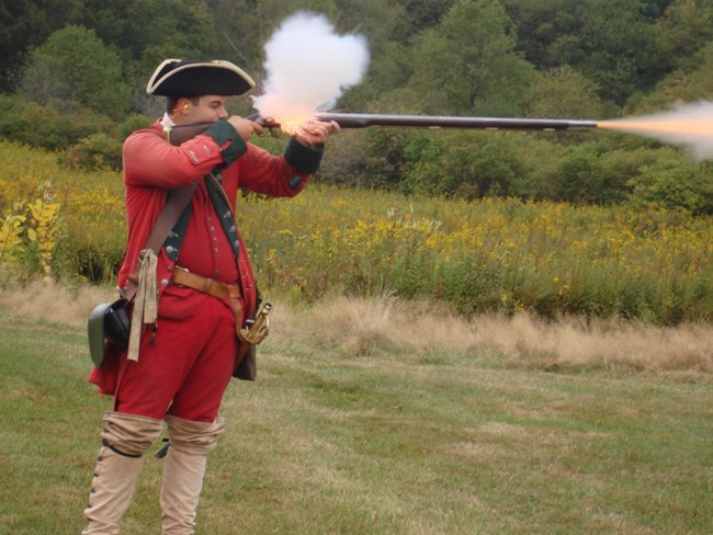 A man in a red uniform fires a black powder musket