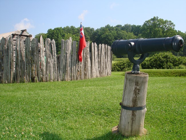 small swivel cannon with Fort Necessity stockade A swivel cannon on a wooden post in the foreground of a fort stockade with green grass and trees.