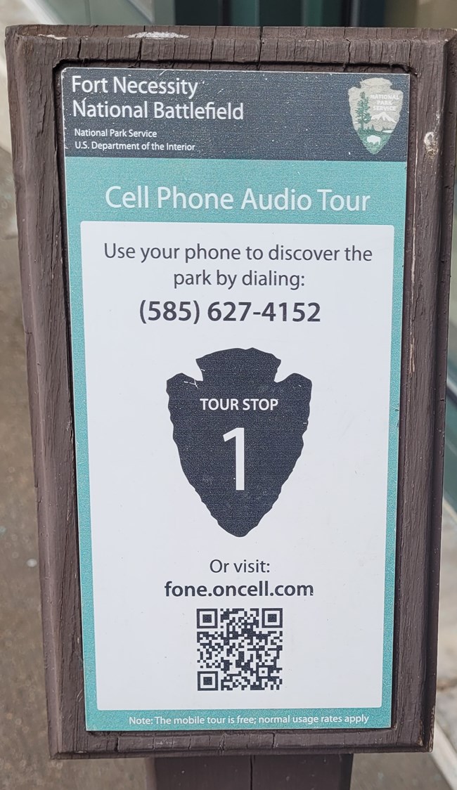 Brown wooden stand with a plaque reading to call 585-627-4152 or scan the QR code for numbered stops