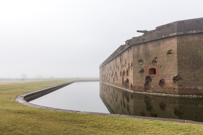 battle-scarred brick walls beside a moat on a foggy morning.