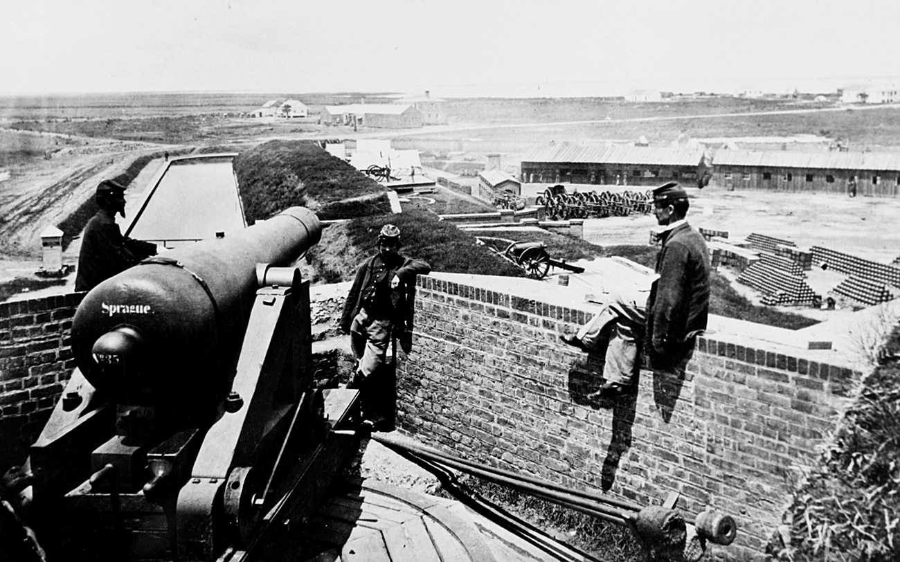 Black and white photograph taken in 1863.Union soldiers stand next to a cannon, and the Workers' Village is visible in the background.