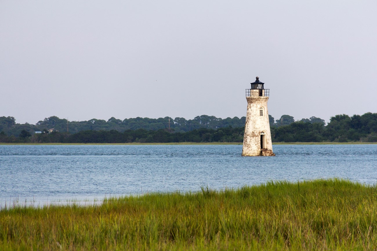 White lighthouse surrounded by water with marsh in the foreground.