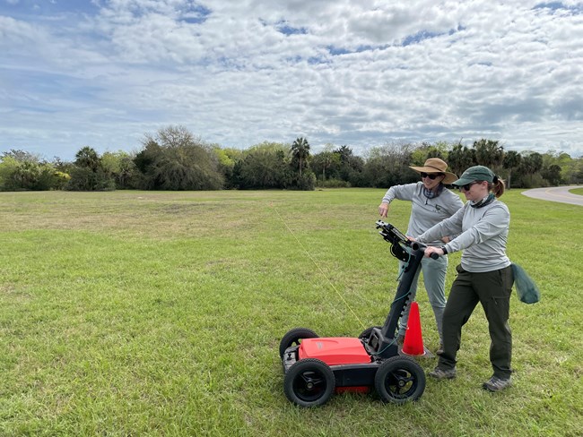 Two archeologists stand in a grassy field with a ground penetrating radar machine.
