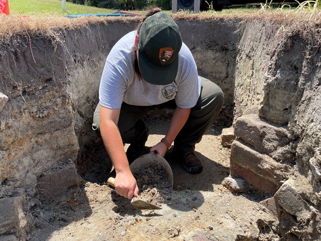 An archeologist kneels while digging in a rectangular hole that is over her head.