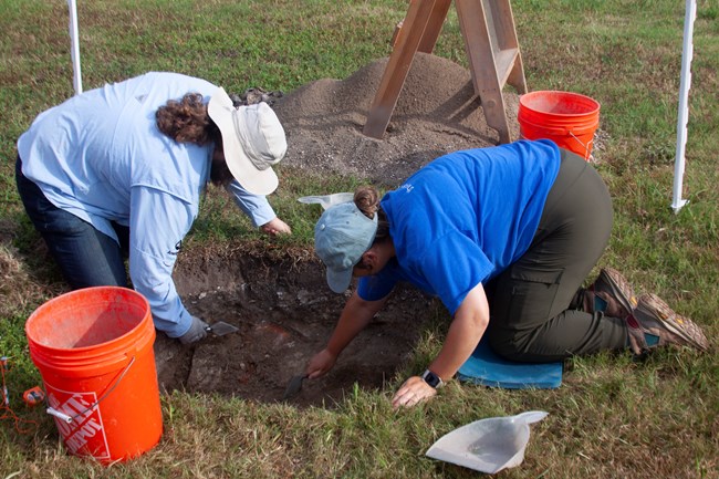 One archeologist digs in a square hole while another archeologist screens the soil nearby.