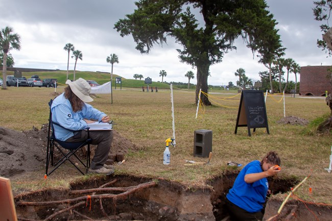 Two archeologists measure and draw the soil layers in a hole.