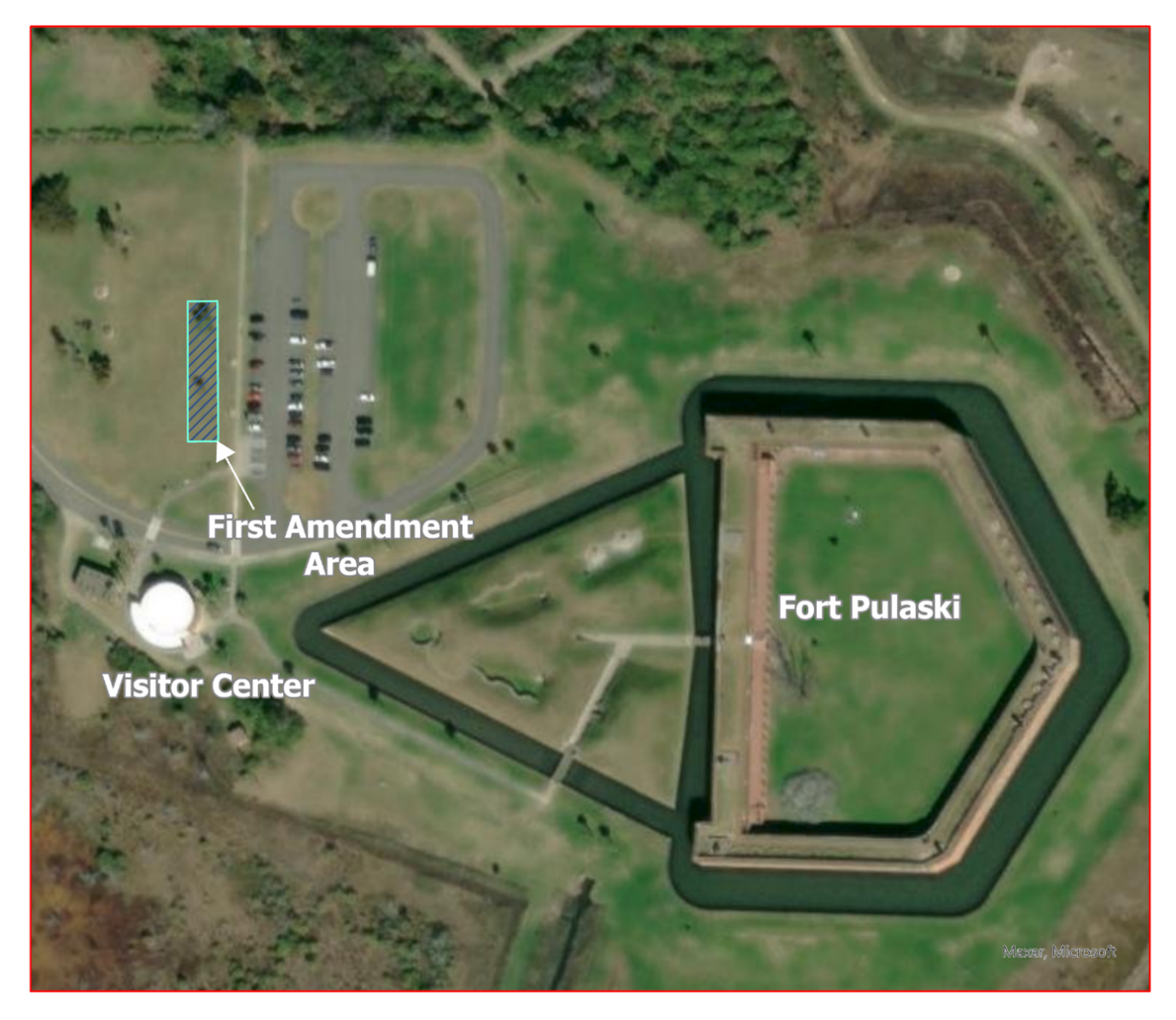 A satellite map that shows the five sided outline of Fort Pulaski, the circlular visitor center, and 2 gray parking lots. to the left of the west parking lot is a small blue box that highlights the first amendment area.
