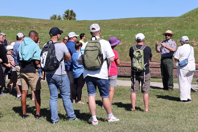 A park ranger gives a talk to a large group of people standing near the moat.