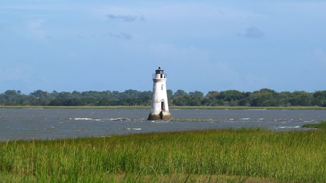 A small, white lighthouse rises from the choppy waters of the Savannah River on a clear, blue day with green, grassy marsh in foreground.