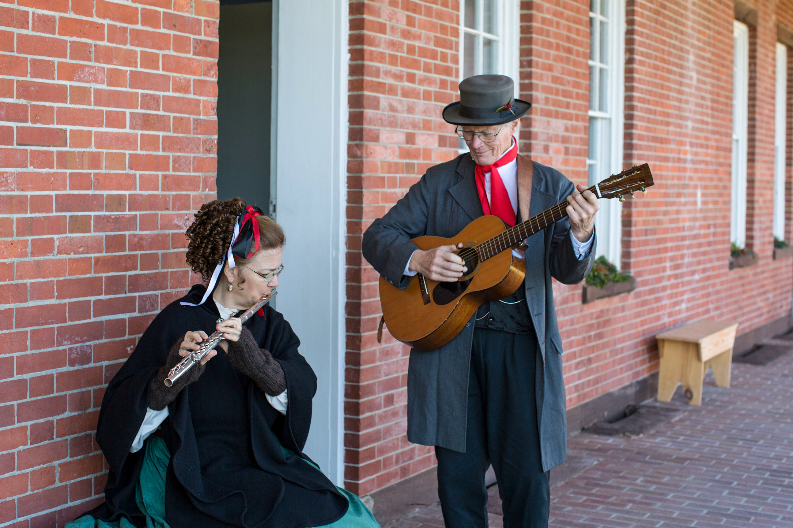 A man dressed in 1800s civilian clothing plays a guitar while standing and a woman dressed in an 1800s dress plays the flute while sitting.