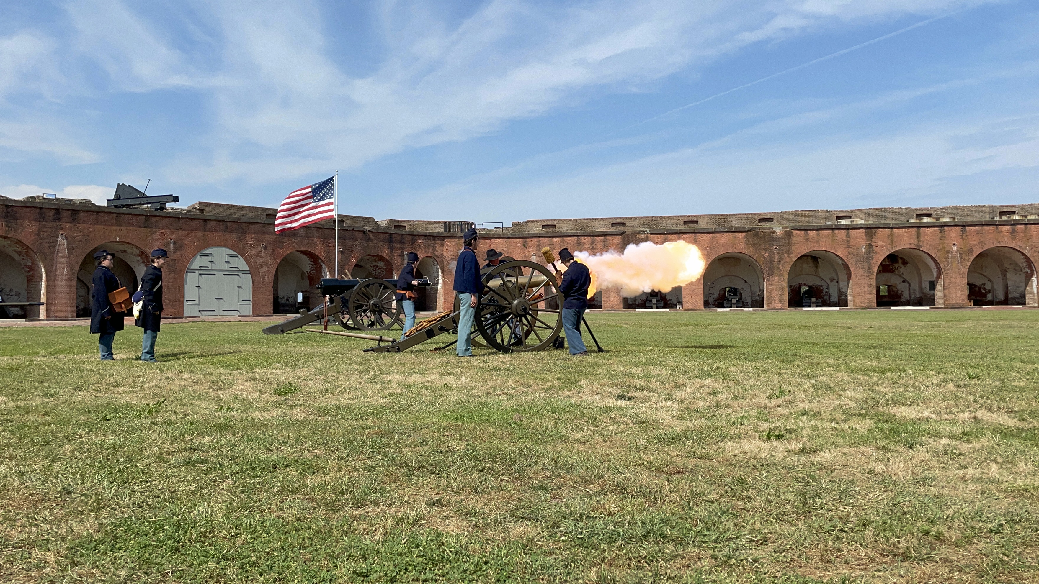 Soliders in blue uniforms surround a small bronze cannon as an explosion of fire erupts from the front of the gun.