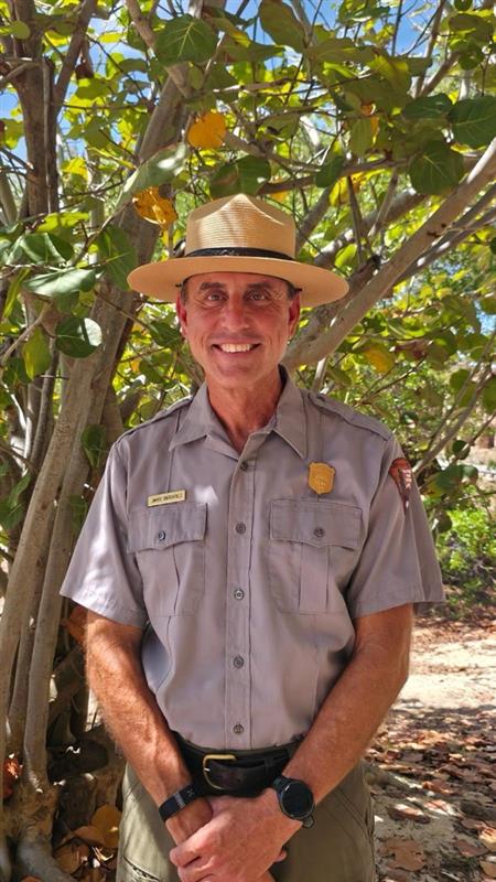 A smiling man, in a green and gray NPS uniform, stands in front of a tree.