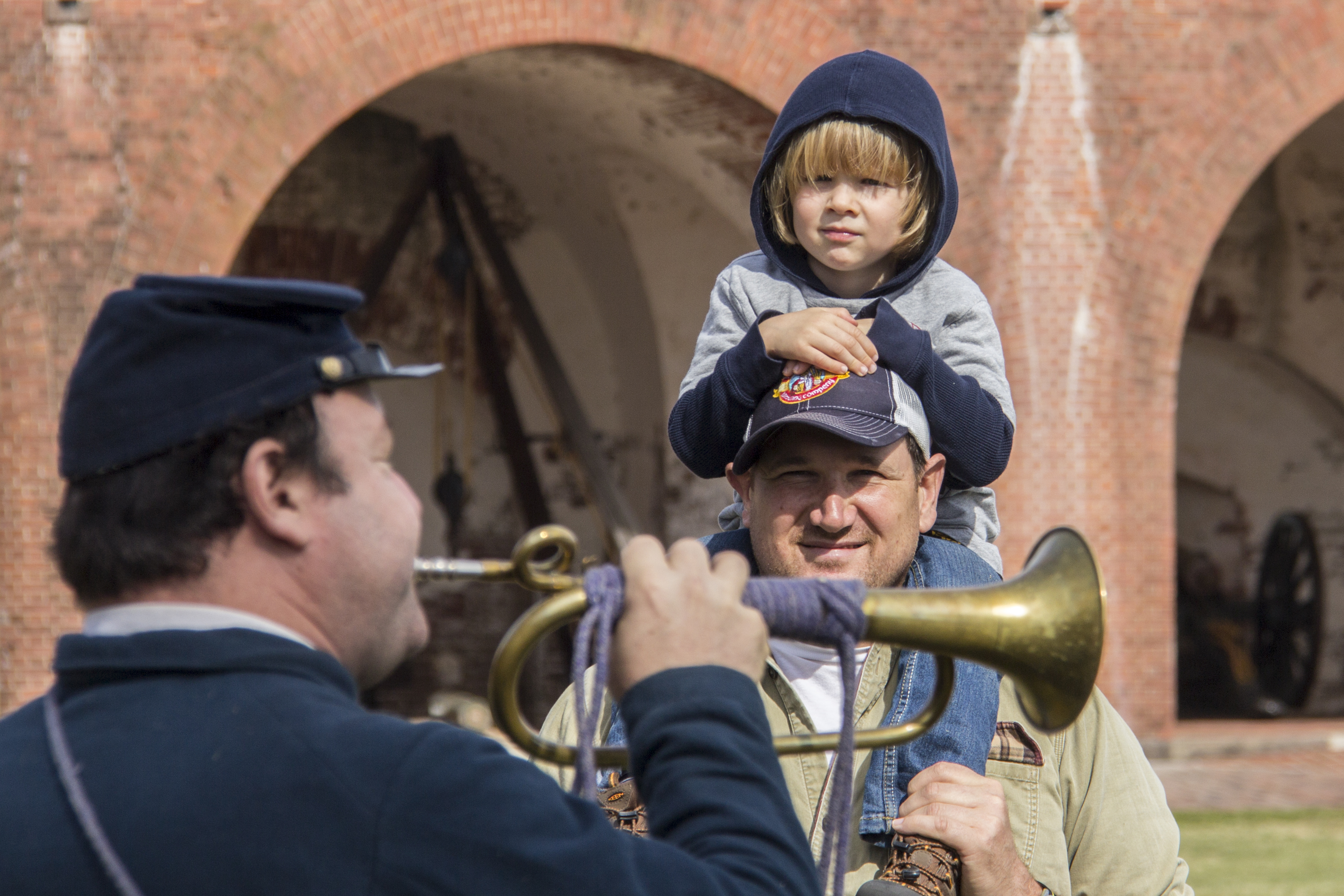 A man in a blue union uniform blows into a brass buglle as a young boy watches as he sits on his father's shoulders.