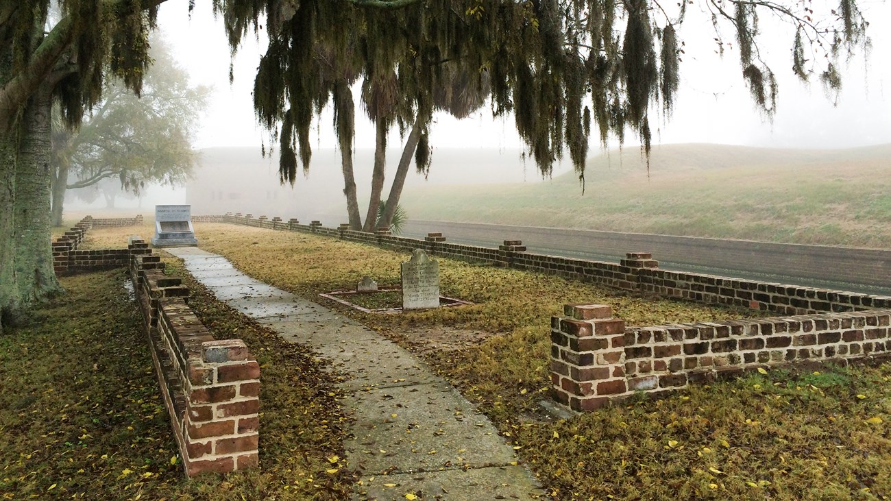 A low rectangular brick wall surrounds a grassy area with fall leaves scattered about. There are two grave stones and a granite memorial inside the area. The walls of a fort are obscured by fog in the background.