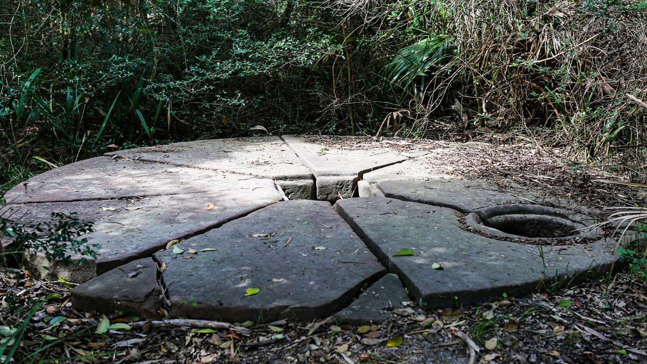 A circular sandstone cistern cap with 2 small openings lays on the ground.