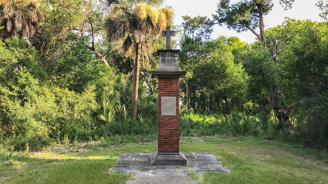 A color image of slender brick column monument with concrete base and and caped with a cross top in a forest like area.