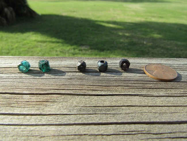 Two green and three black glass faceted beads on a wooden board. Beads are approximately one quarter of an inch in diameter and length.