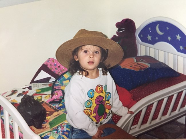 Young child sitting on a bed wearing a Park Ranger hat.