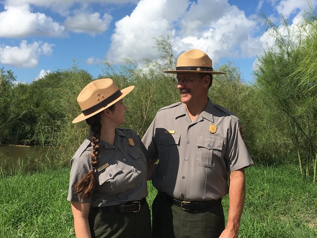 Two rangers stand in uniform smiling at each other. Background is green grass and trees with cloudy blue sky.