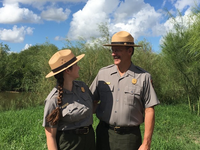 My Park Story - Lauren (2) Two rangers stand in uniform smiling at each other. Background is green grass and trees with cloudy blue sky.
