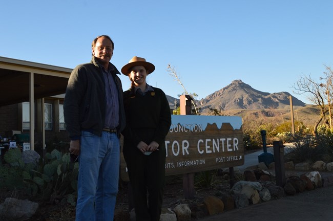 Ranger stands with her father in front of Visitor Center sign with mountain in background.
