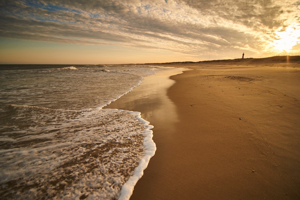 Waves flow against a peaceful beach with the top of a lighthouse seen in the distance