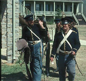 Soldiers in fatigue uniforms walk the grounds of Fort Scott carrying equipment