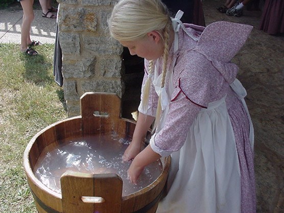 A young woman in a prairie dress and apron washes clothes in a wooden bucket