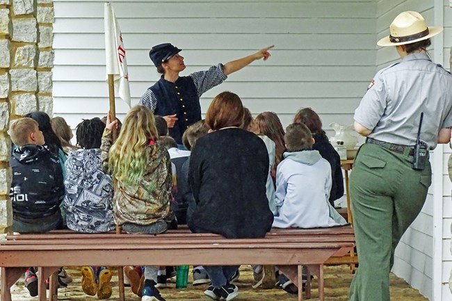 An interpreter in period dress gives a lesson to schoolchildren at a station as part of the Life on the Frontier program as a uniformed ranger watches.