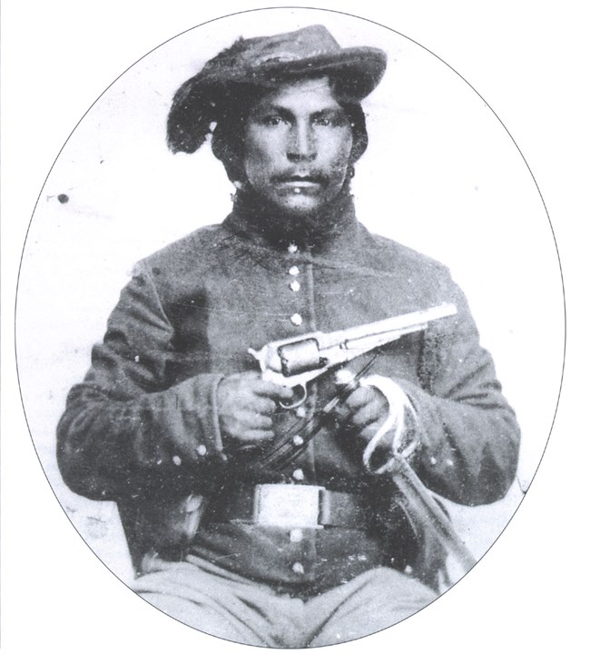 Black and white photo of a Native American in soldier uniform holding a gun and sword.