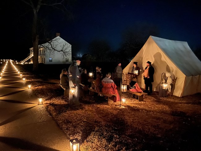 Image of living history volunteers outside of a tent lit by candles.