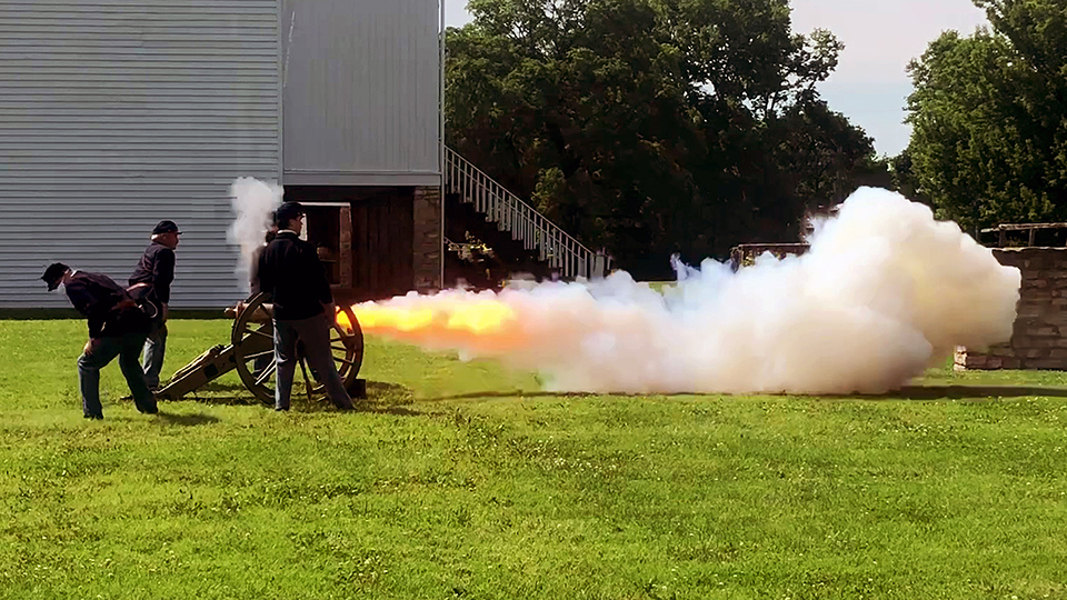 Four people dressed in Civil War uniforms firing a 12# Mountain Howitzer. Smoke and flames are coming out of the barrel.