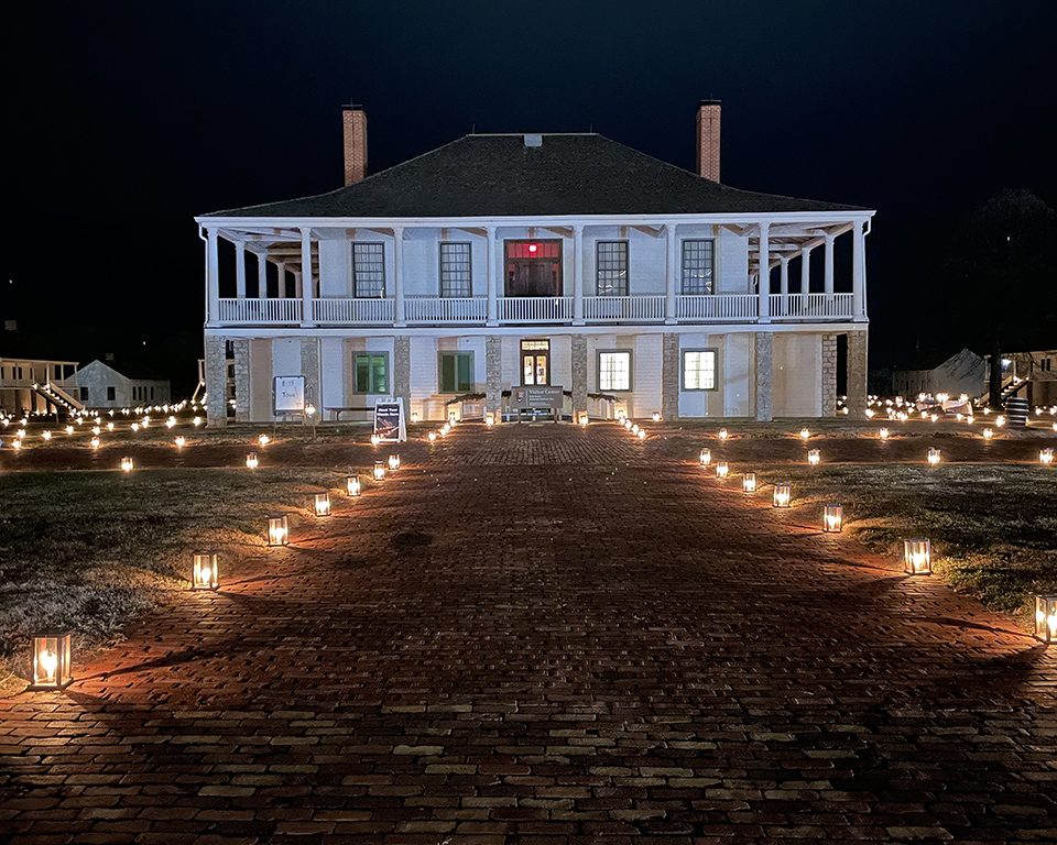 Post Hospital during the Candlelight Tour. Image of a white building with stone pillars on the first floor and a white railing around the porch on the second. Candle lanterns line the sidewalk leading to the building.