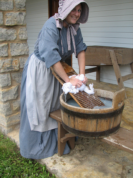 1840s Laundress Woman dressed in a blue and white checked dress and bonnet washing close with a bucket and wash board.