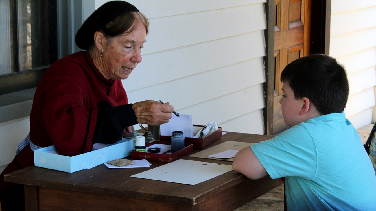 Female reenactor with young visitor using an inkwell. Female reenactor with young visitor using an inkwell.