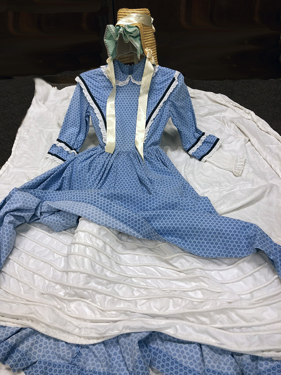 Image of a straw bonnet, blue dress, and a white petticoat laid out on the floor. Image of a straw bonnet, blue dress, and a white petticoat laid out on the floor.