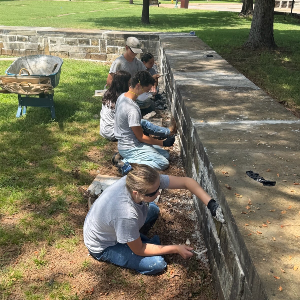 A group of five young people wearing YCC uniform shirts sitting on the ground, repairing the mortar on a low stone wall.