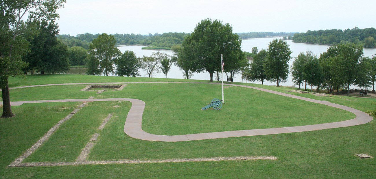 A sidewalk winds through gray stone lines in grass with tress and a river in the background.