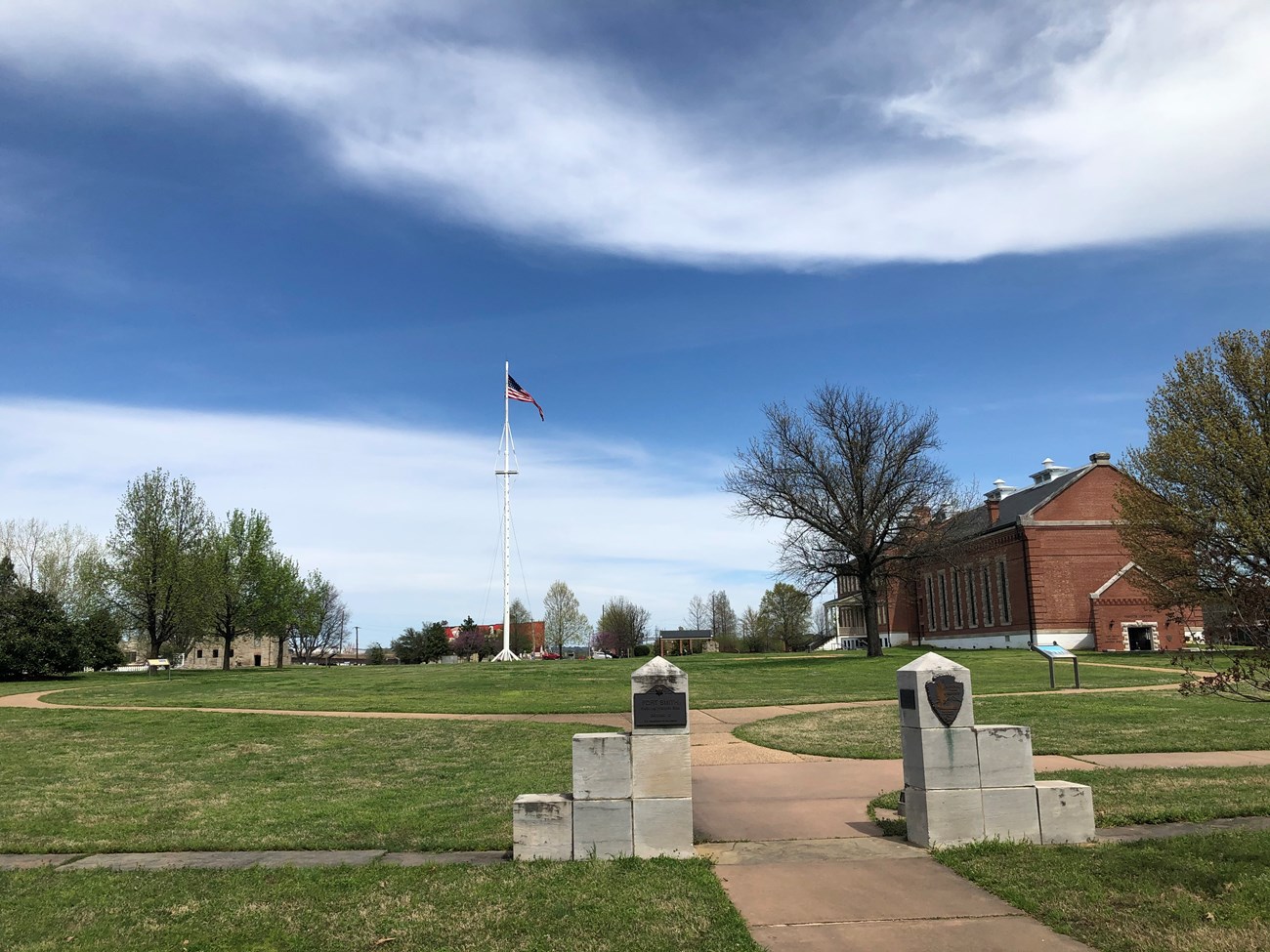 Two tiered gray stone markers form an opening on a sidewalk in the foreground with an open green lawn, flag pole, and red brick building in the back ground on a sunny day.