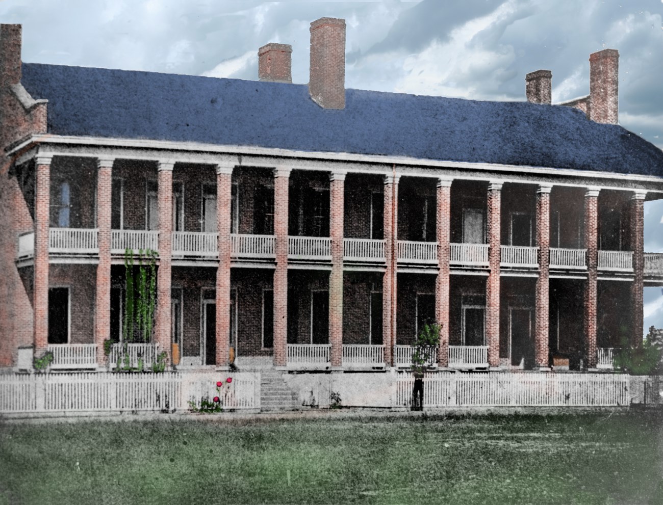Colorized historic photo of two story red brick building with balcony and porch spanning the length of the building.