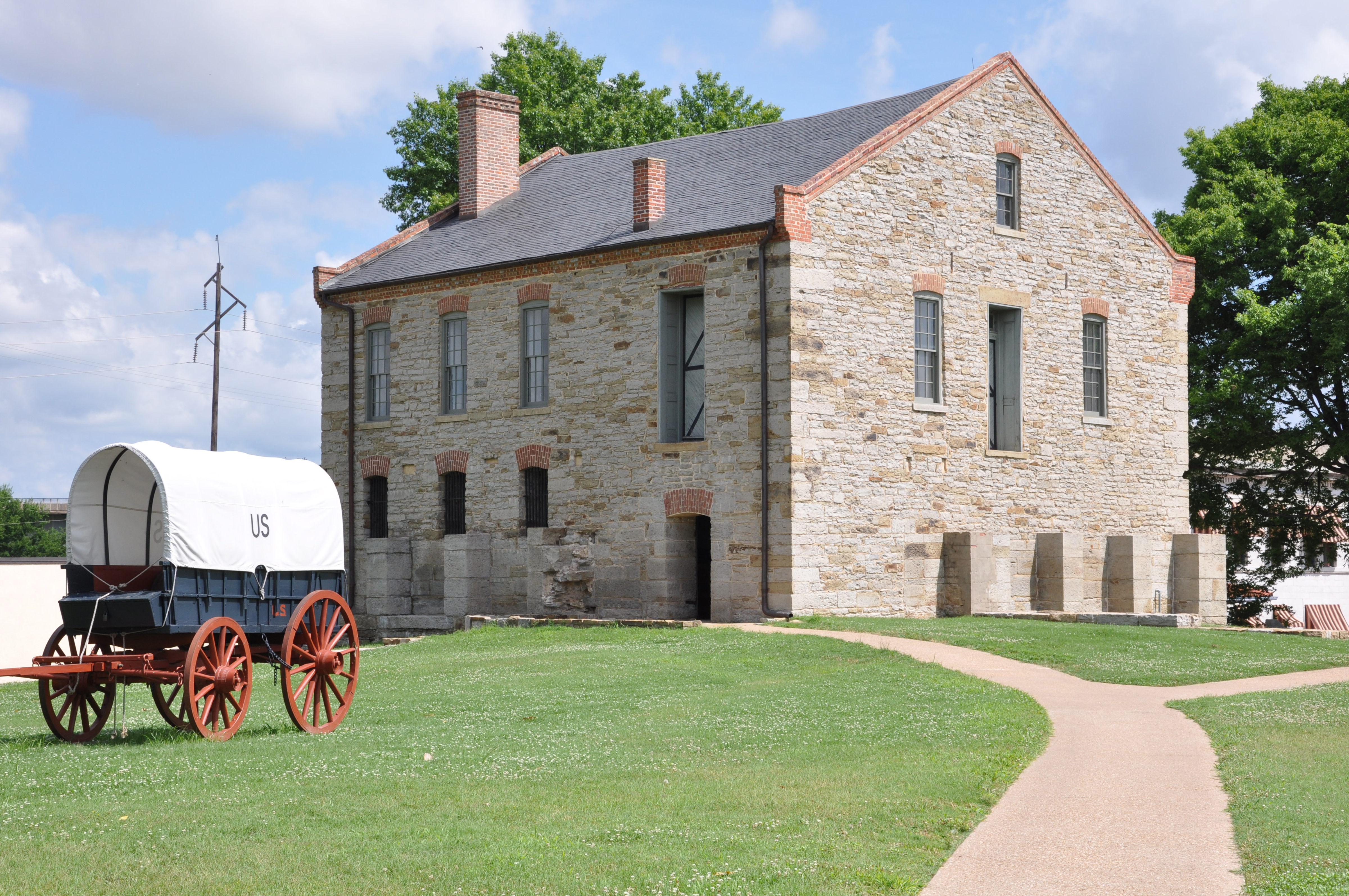 An Army supply wagon with red wheels, navy blue body, and white canvas cover stands in front of a two story tan stone building, the commissary, on a sunny day.