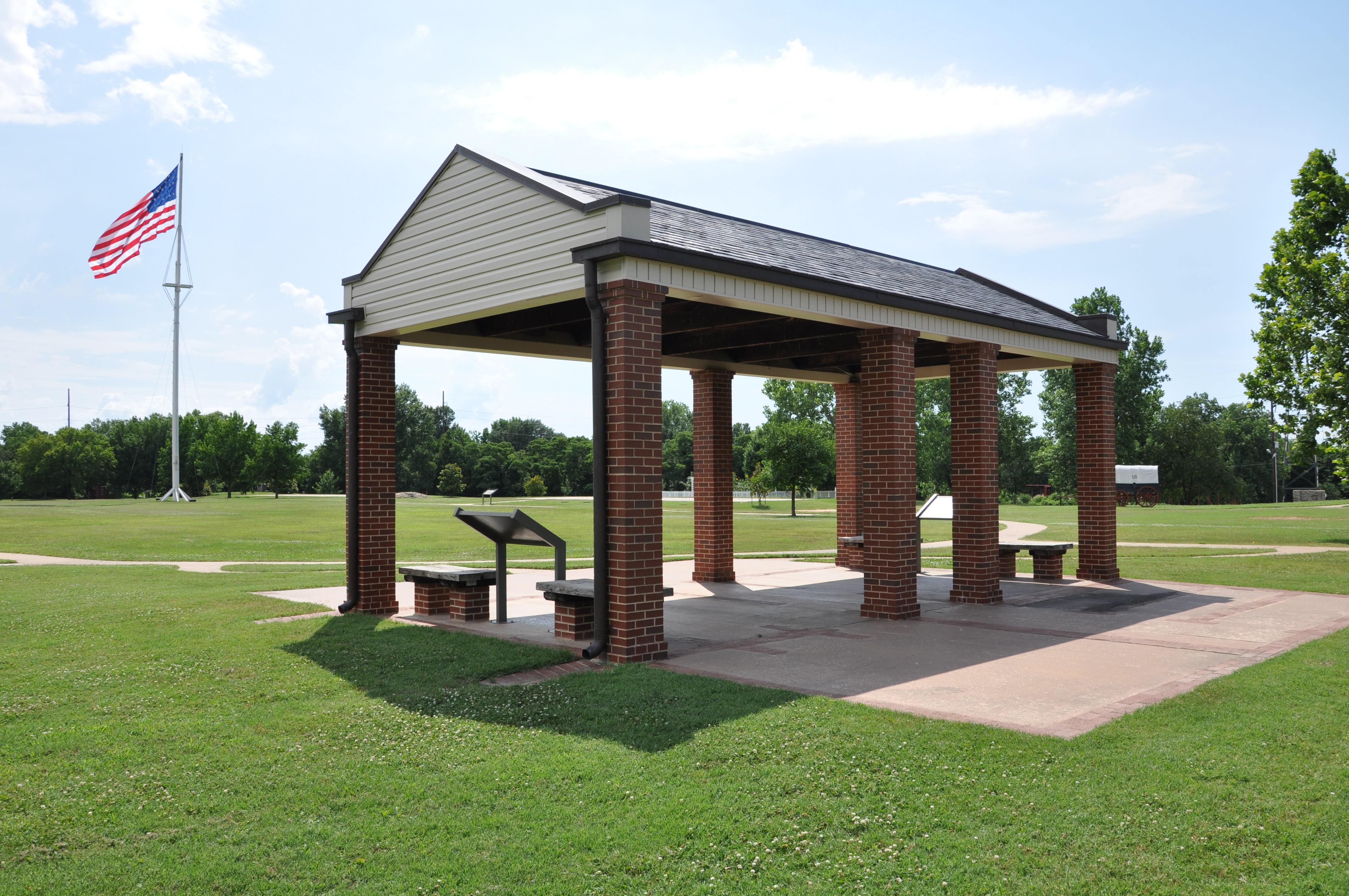 A red brick rectangular gazebo with US flag flying in the background on a sunny day.