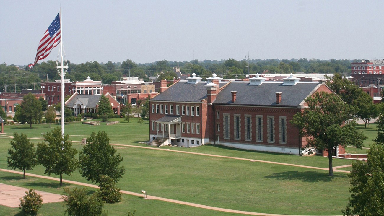 Aerial view of a large 37 star flag flying on a 100 foot tall flag pole in front of the red brick visitor center on a clear sunny day