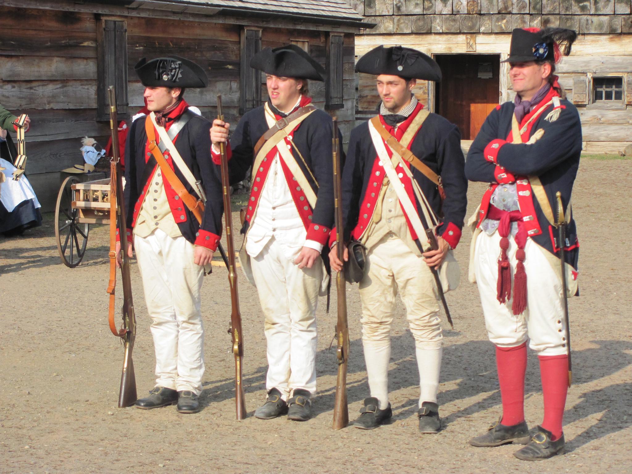 Four Continental Soldiers stand shoulder to shoulder. One has his arms crossed, the other three hold muskets by their sides.