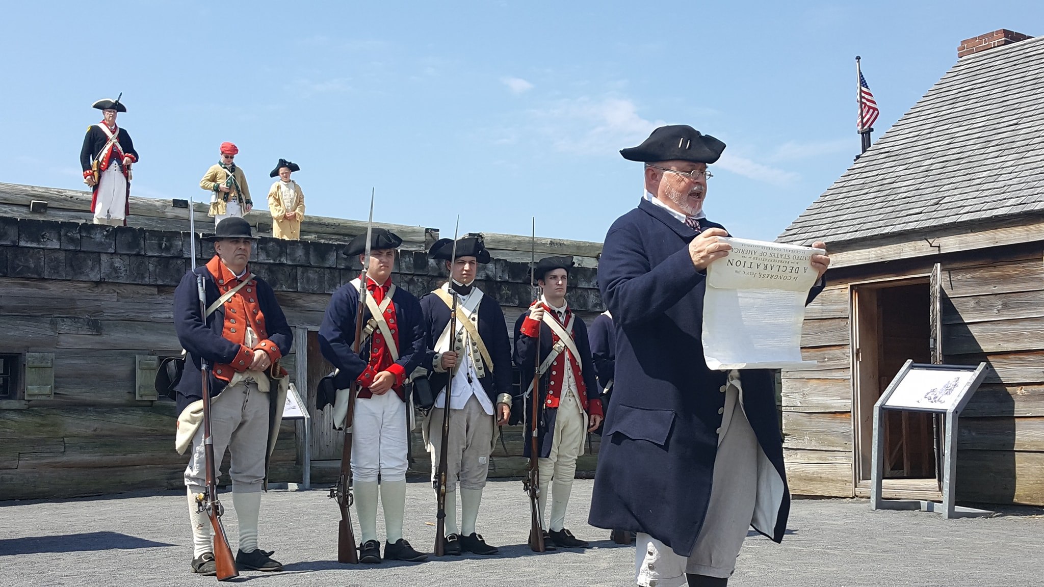 A row of Continental soldiers stand in the fort. A step in front is a man in a wool jacket holding and reading a large sheet of parchment paper. 