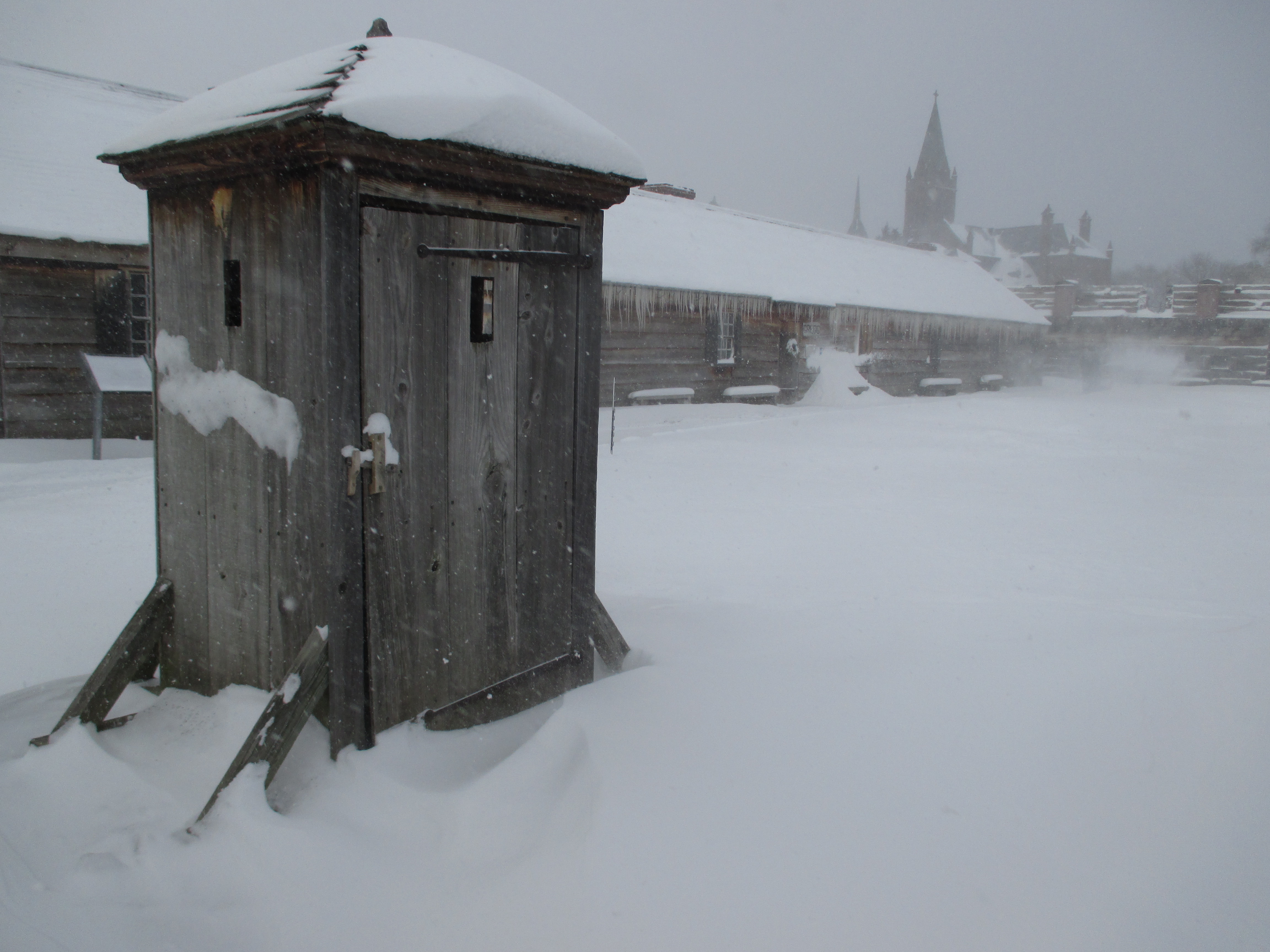 A view of a cold, windy open area with snow blowing all around. The sun is muted under the dark clouds above.
