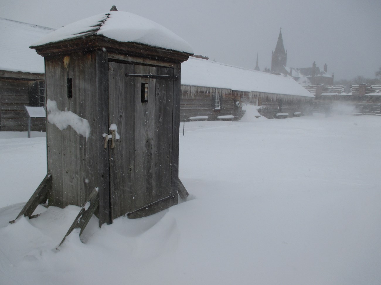 A view of a cold, windy open area with snow blowing all around. The sun is muted under the dark clouds above.