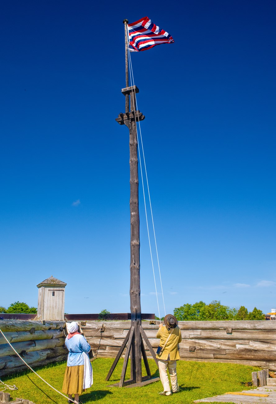 A man in and woman in 18th C clothing stand below a striped flag while pulling it to the top of the flag pole.